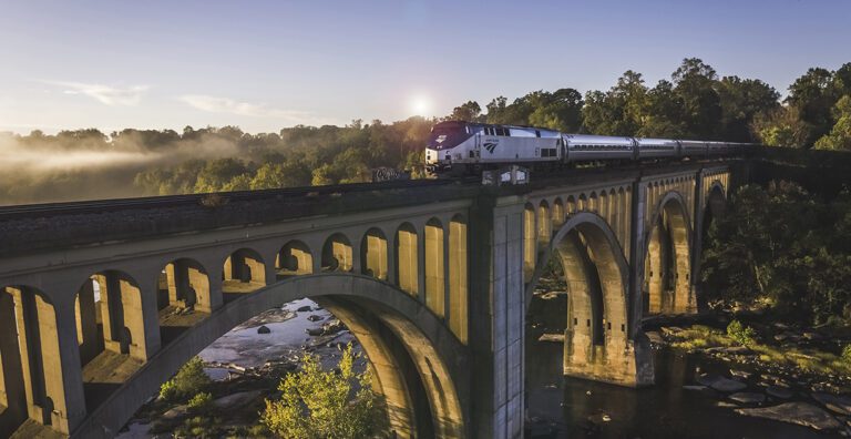 Amtrak train crossing a bridge over a river with the sun shining behind the trees